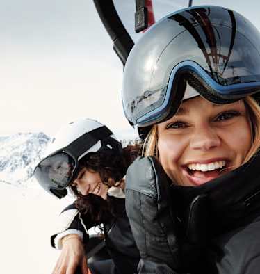 Eine Frau mit Skibrille und Helm auf dem Kopf sitz in einem Sessellift und lacht uns entgegen. Neben ihr sitzt eine weitere lächelnde Frau.