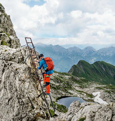Ein Kletterer nimmt die Stahlleiter, im Hintergrund ist ein Bergsee zu sehen