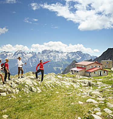 Vier Wanderer schauen in die Ferne. Im Hintergrund eine Berghütte und eine Gipfelkette.