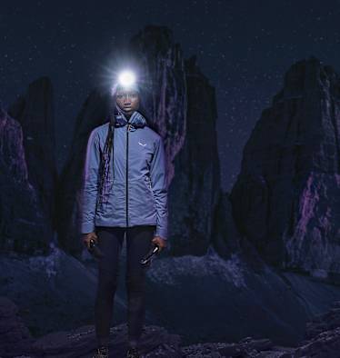 Frau mit Stirnlampe in Speedhiking Outfit von Salewa in der Nacht, im Hintergrund Berge in der Dunkelheit.