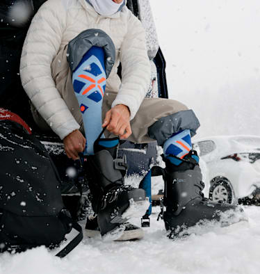 Ein Skifahrer sitzt am Parkplatz und zieht sich im Schnee seine Skischuhe an.