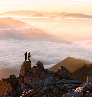 Zwei Wanderer auf einem Felsen im Hochgebirge überblicken das von Wolken bedeckte Tal.