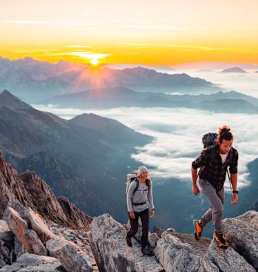 Zwei Wanderer steigen bei Sonnenaufgang auf einen Dolomitengipfel im Trentino. Im Tal ein Nebelmeer.