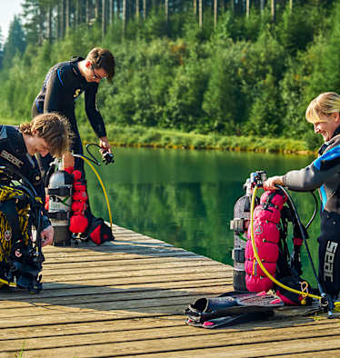 Drei Taucher in Neoprenanzügen knien auf einem Holzsteg am Grüblsee
