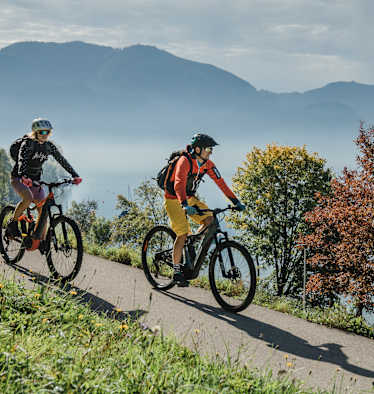 Zwei Radfahrer auf einer Straße in Oberösterreich, im Hintergrund Berge und buntgefärbte Bäume.
