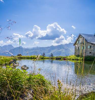 Zwei Mountainbiker radeln an einem Bergsee entlang. Daneben steht eine Hütte.