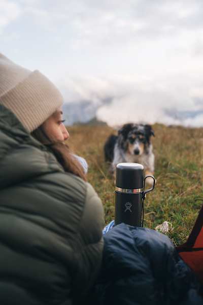 Frau mit Hund bei einer Pause in den Bergen, neben einer schwarzen Hydro Flask Hot Flask & Cup, umgeben von nebliger Landschaft.