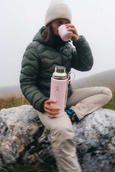 Frau in Winterkleidung sitzt auf einem Felsen und trinkt ein heißes Getränk aus der rosa Hydro Flask Hot Flask & Cup in nebliger Natur.