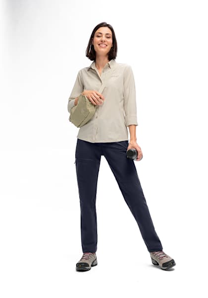 Frau in Maier Sports Outdoorbekleidung mit nachhaltiger Wanderhose und beigem Funktionshemd, kombiniert mit Wanderschuhen und Umhängetasche, vor weißem Hintergrund.