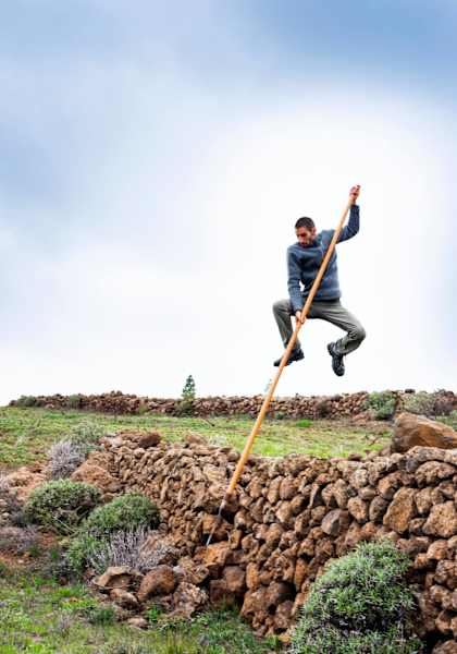 Ein junger Mann springt mit Hilfe eines Holzstabes über eine alte Mauer