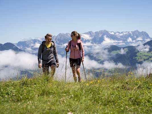 Ein Mann und eine Frau wandern über eine Wiese, hinter ihnen ein Bergpanorama.