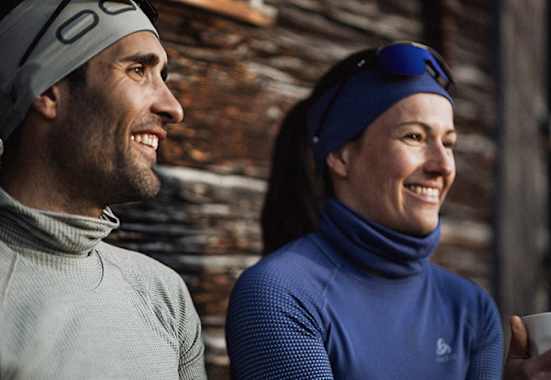 Zwei Personen in funktionaler Winterbekleidung von Odlo, genießen eine Pause im Freien vor einer rustikalen Holzhütte.