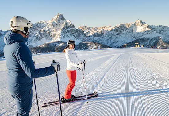 zwei skifahrer stehen auf der piste und schauen sich an, der himmel ist strahlend blau und die schneebedeckten bergspitzen stehen im hintergrund