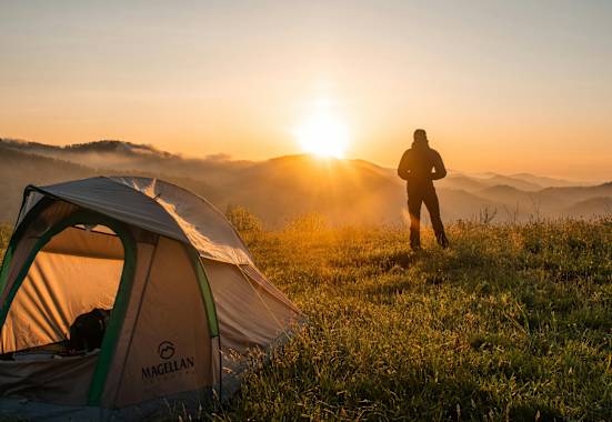 Ein Mann steht vor seinem Zelt und schaut zu wie die Sonne hinter den Bergen untergeht.