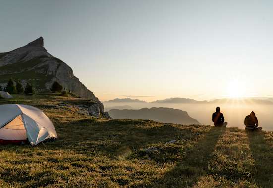 Zwei Personen sitzen vor ihrem Zelt und betrachten den Sonnenuntergang vor einer beleuchteten Bergkulisse.