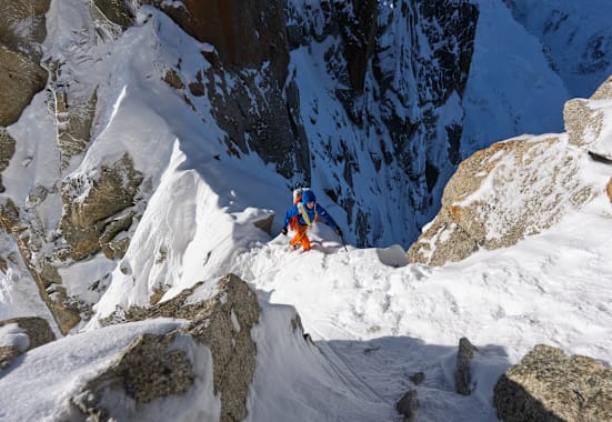 Ein Bergsteiger mit blauer Jacke und orangefarbener Hose erklimmt einen Grat. Dabei ist er mit einem Seil gesichert.