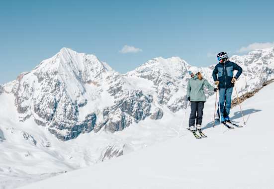 Ein Skifahrer und eine Skifahrerin stehen auf einem Hang und genießen das verschneite Bergpanorama im Ortlergebiet.