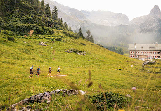 Vier Trailrunner laufen entlang einer Wiese auf einem Berg in Richtung einer Hütte mit roten Fensterläden.