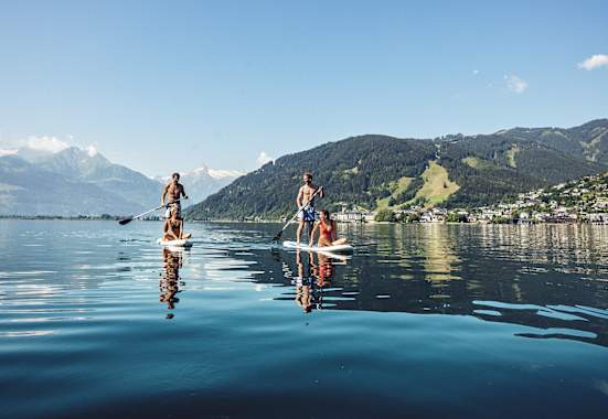 Zwei Männer tauche mit einem Puddle ihr Stand-up-paddeln an. Jeweils eine Frau, sitz am vorderen Ende. Sie gleiten über denn Zeller See. Hinter ihnen ein Bergpanorama.