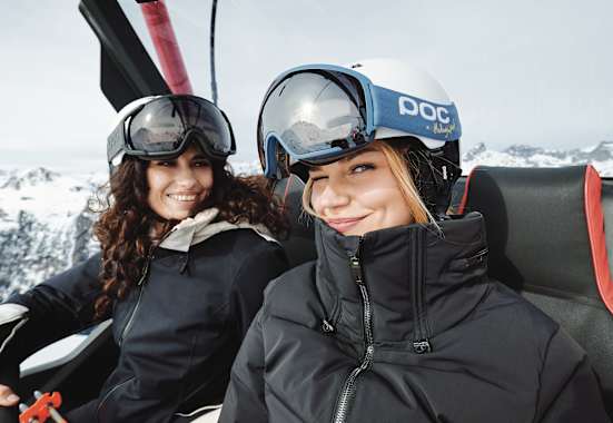 Eine blonde Frau zwinkert in die Kamera. Eine andere Frau, die neben ihr sitzt lächelt ihr dabei zu.