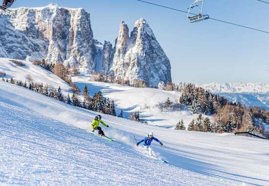 Zwei Skifahrer fahren eine frisch präparierte Piste in der Dolomitenregion Seiser Alm hinunter. Im Hintergrund ist das verschneite Bergpanorama zu sehen.