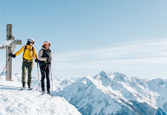 Eine Frau und ein Mann stehen neben dem Gipfelkreuz auf Tourenskiern und blicken über die verschneite Berglandschaft.