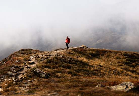 Person wandert in roter Outdoor-Jacke auf einem Bergpfad durch eine neblige Gebirgslandschaft.