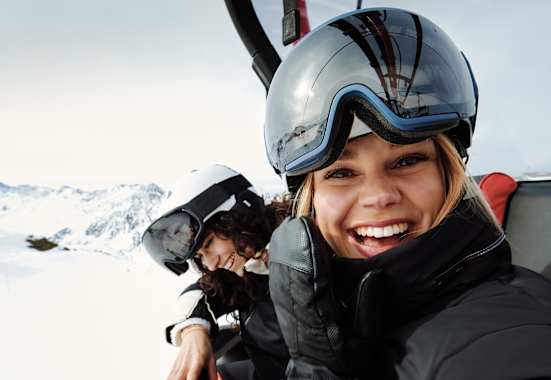 Eine Frau mit Skibrille und Helm auf dem Kopf sitz in einem Sessellift und lacht uns entgegen. Neben ihr sitzt eine weitere lächelnde Frau.
