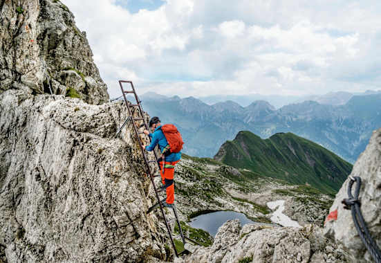 Ein Kletterer nimmt die Stahlleiter, im Hintergrund ist ein Bergsee zu sehen