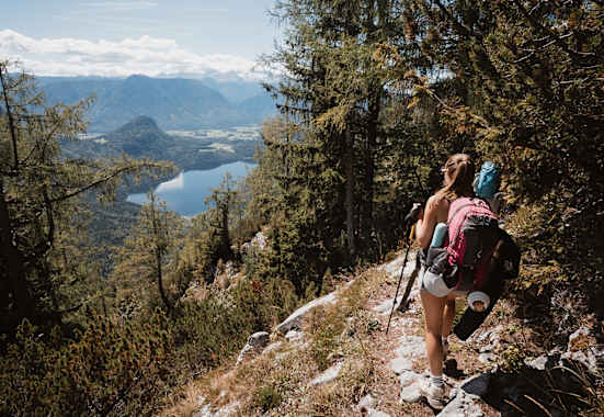 Eine Wandrerin mit Rucksack und Ausrüstung, die durch den Wald auf den Altauseer See hinunterblickt.