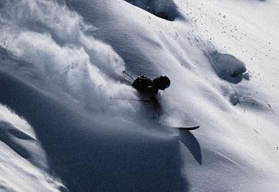 Skifahrer gleitet dynamisch durch unberührten Tiefschnee, wirbelt Pulverschnee auf und meistert eine steile Abfahrt in alpiner Winterlandschaft.