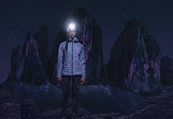 Frau mit Stirnlampe in Speedhiking Outfit von Salewa in der Nacht, im Hintergrund Berge in der Dunkelheit.