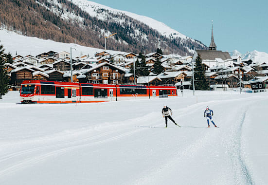 Ein Tal mit 2 Langläufern, einer Bahn und verschneite Tannen.