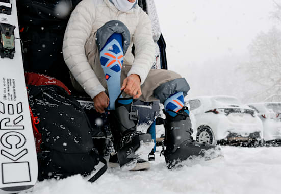 Ein Skifahrer sitzt am Parkplatz und zieht sich im Schnee seine Skischuhe an.