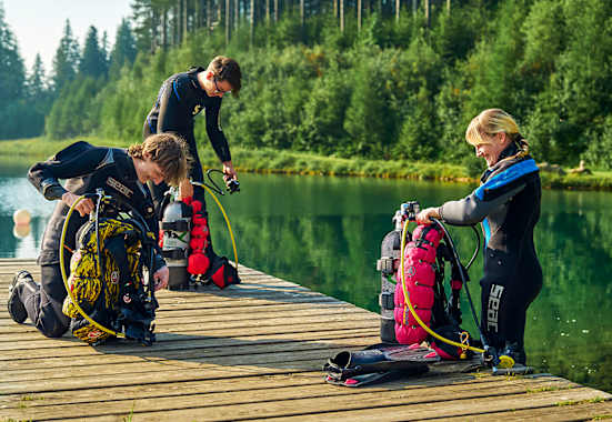 Drei Taucher in Neoprenanzügen knien auf einem Holzsteg am Grüblsee