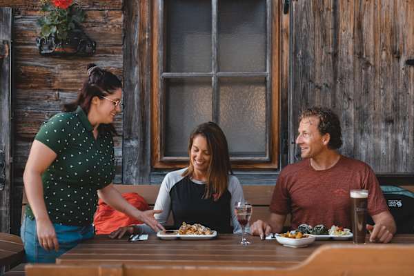 Zwei Personen sitzen am Tisch vor der Holzalm im Alpbachtal und bekommen ihr Essen serviert: Spinatknödel und Kaiserschmarrn.