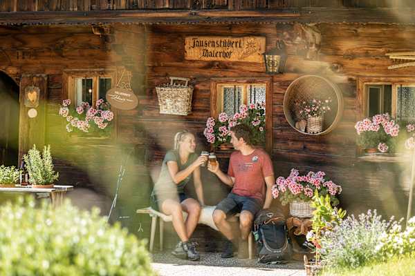 Zwei Personen sitzen vor der urigen Hauswand der Jausenstation Oberthaler in der Ferienregion Alpbachal und stoßen mit einem Bier an.