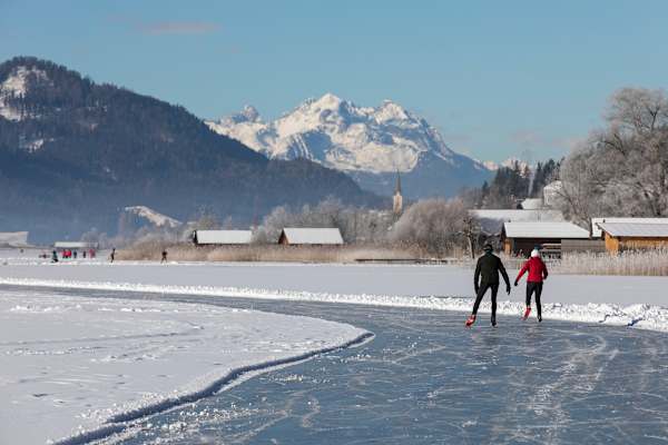 Zwei Schlittschuhläufer auf der präparierten Loipe auf dem Weißensee mit Bergpanorama im Hintergrund