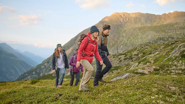 Eine Gruppe von Wanderern in funktionaler Outdoor-Bekleidung bewegt sich durch eine malerische Berglandschaft. Grüne Wiesen und majestätische Gipfel bilden eine beeindruckende Kulisse.