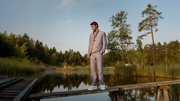 Ein Mann in modischer Kleidung in matten Farbtönen auf einem Holzpfad umrandet von einem See und Wäldern.