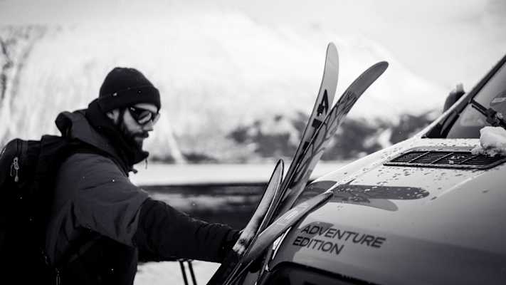 Person lädt Skier auf einen Camper vor schneebedeckter Bergkulisse in Nord-Norwegen.
