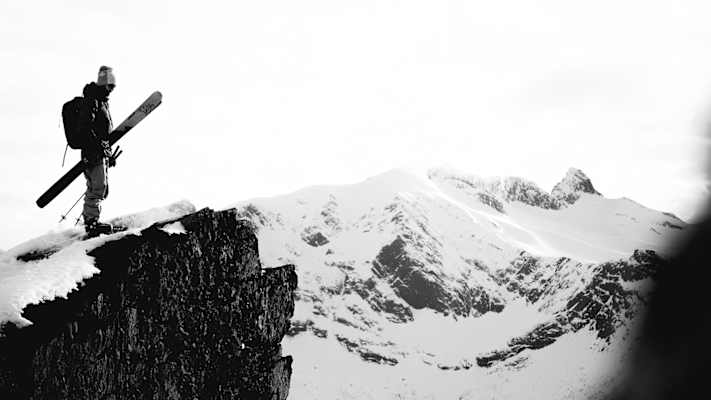 Skitourengeher steht mit Skiern auf einer verschneiten Felskante, vor den schneebedeckten Gipfeln der Lyngenalpen in Nord-Norwegen.