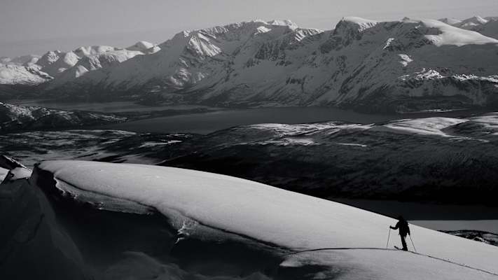 Skitourengeher auf einer verschneiten Bergflanke mit Panoramablick auf Fjorde und die schneebedeckten Lyngenalpen in Nord-Norwegen.