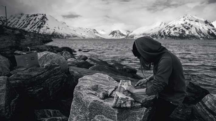 Person filetiert einen Fisch auf Felsen am Fjord, umgeben von schneebedeckten Bergen in Nord-Norwegen.