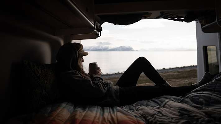 Person liegt entspannt im Camper und blickt auf einen Fjord mit schneebedeckten Bergen in Nord-Norwegen.