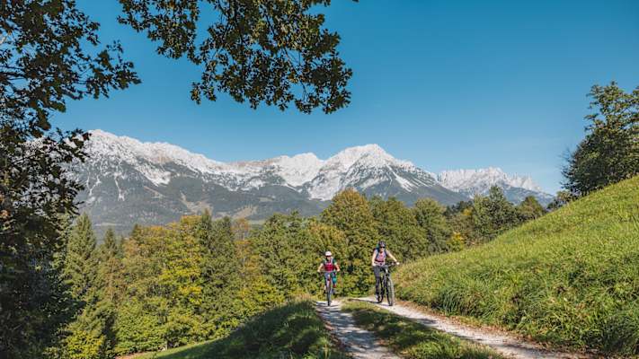 Ein Mann und eine Frau radeln auf einem Forstweg bergauf. Im Hintergrund stehen schneebedeckte Berge.