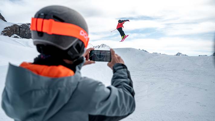 Ein Skifahrer macht einen Stunt über einen kleinen Schneehügel, während ihn ein anderer Skifahrer dabei filmt.