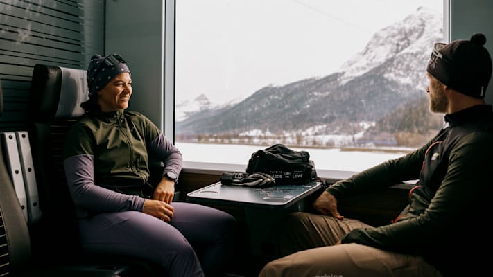 Ein Paar sitzt im Zug mit Bergpanorama und lächelt sich an.