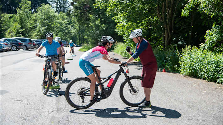 Mountainbike-Guide Georg Fröhlich zeigt einer Fahrerin beim Techniktraining im Freien die richtige Position auf dem Bike, weitere Teilnehmer:innen warten im Hintergrund.