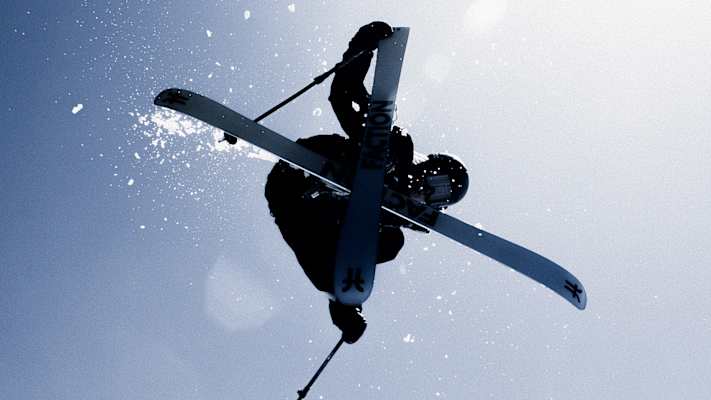 Freestyle-Skifahrer in der Luft bei einem Trick, mit Sicherheitsausrüstung und Ski unter blauem Himmel.